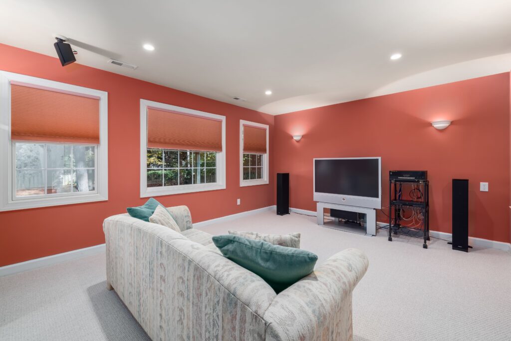 Spacious home theater room with orange walls and a comfortable couch, featuring a flat-screen TV and ample natural light.